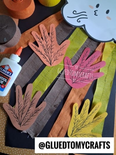Colored construction paper turkey feet, glue, and strips of orange, yellow, and brown paper are arranged on a table, with a cloud cutout at the top. The handle @GLUEDTOMYCRAFTS is displayed at the bottom.