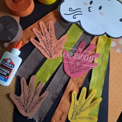Fall craft supplies, including paper leaves, a cloud with a face, streamers, glue, and colored paper, arranged on a table. @GLUEDTOMYCRAFTS watermark is visible in the image.
