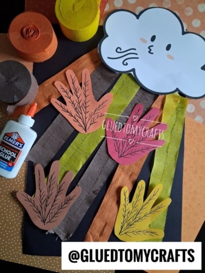 Fall craft supplies, including paper leaves, a cloud with a face, streamers, glue, and colored paper, arranged on a table. @GLUEDTOMYCRAFTS watermark is visible in the image.