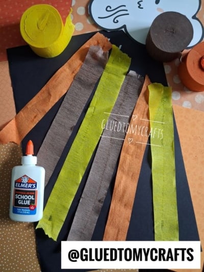 Black paper with glue, torn strips of orange, brown, and yellow tissue, a white school glue bottle, and rolls of crepe paper, arranged for a craft. @GLUETOMYCRAFTS is noted at the bottom.