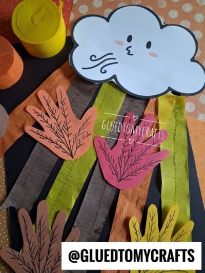 A paper craft featuring a smiling cloud blowing wind, with colored paper strips and handprint cutouts below, creating a fall or autumn scene. @GLUETOMYCRAFTS appears at the bottom.
