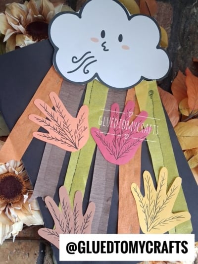 A blowing cloud craft showing a smiling cloud with rain made from colorful handprint cutouts and brown, orange, and yellow strips, surrounded by artificial sunflowers and autumn leaves. @GLUEDTOMYCRAFTS watermark is visible.