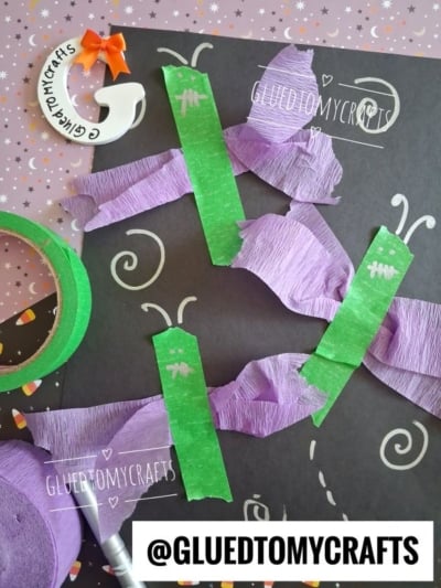 Four purple and green caterpillar crafts made from crepe paper and tape are arranged on a black sheet with doodles. A green tape roll, star-patterned background, and @GLUEDTOMYCRAFTS text are also visible.