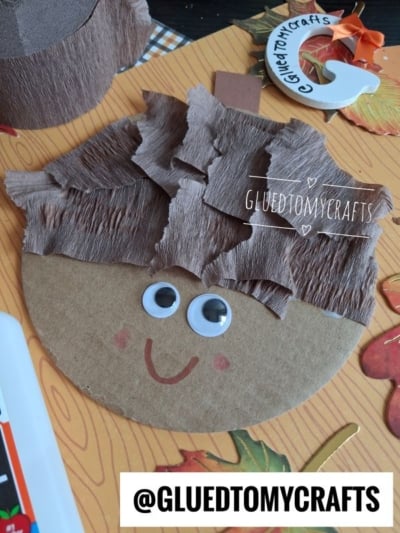 A round cardboard face with googly eyes, a drawn smile, and layered brown crepe paper hair, lying on a decorated orange surface with craft supplies around it. @GLUEDTOMYCRAFTS is visible on the image.