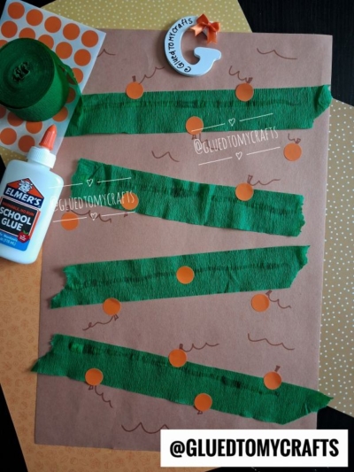 Brown paper with green crepe paper strips, orange dot stickers, wavy marker lines, a bottle of Elmers glue, green ribbon, a pumpkin craft, and a polka dot sheet—this festive setup is perfect for FALL CRAFTS FOR KIDS. Watermarks say @gluedtomycrafts.