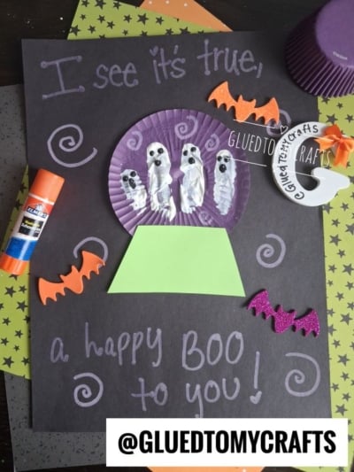 This fun Halloween craft for kids features white ghost fingerprints on a purple cupcake liner, surrounded by bats, swirls, and glitter accents on black paper. Text says I see it’s true, a happy boo to you!.