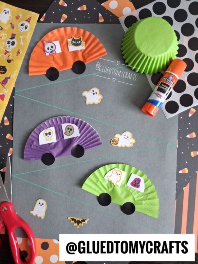 Halloween Crafts for Kids: Halloween craft scene with cupcake liners made into cars, decorated with spooky stickers on gray paper. Scissors, glue stick, and Halloween-themed papers and stickers are also visible. GLUEDTOMYCRAFTS text at bottom.