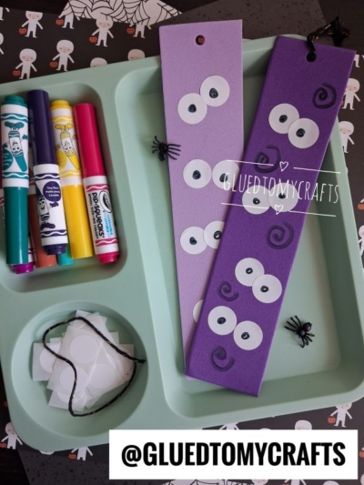 DIY Halloween bookmarks displayed in a mint-green tray, featuring two purple foam bookmarks decorated with multiple paper eyes, alongside markers, white circle stickers, string, and small toy spiders.