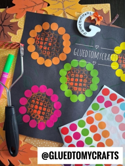 A fall-themed craft scene with colorful dot stickers forming flower shapes on black paper, a craft roller, patterned leaves, and a GLUED TO MY CRAFTS tag near the center, perfectly capturing the creativity of a potato masher stamped craft.