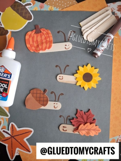 Four craft stick bugs with fall-themed decorations—pumpkin, sunflower, acorn, and leaf—are arranged on colored paper. Supplies like glue, markers, and paper are nearby for easy crafting. This charming scene is a perfect example of FALL CRAFTS FOR KIDS. @GLUEDTOMYCRAFTS is written on the image.