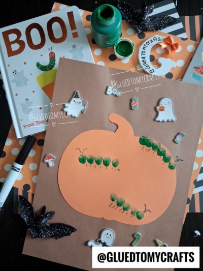 A festive brown paper features an orange pumpkin cutout, green fingerprint caterpillars, and Halloween stickers—one of many fun FALL CRAFTS FOR KIDS. Nearby are a BOO! book, paint, a marker, and black glitter bats set against a playful polka-dot background.