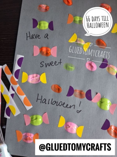 A children’s Halloween craft on dark gray paper features colorful thumbprint “candies” made with painted fingerprints and paper wrapper ends, surrounding the handwritten message “Have a Sweet Halloween!”