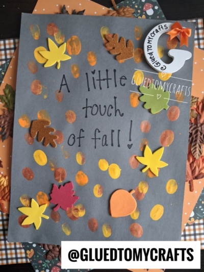 A dark paper with A little touch of fall! written on it, decorated with foam autumn leaves and orange/yellow paint dots. A sticker reads @GLUEDTOMYCRAFTS. Fall-themed paper is visible underneath—perfect inspiration if you’re searching for fun FALL CRAFTS FOR KIDS.