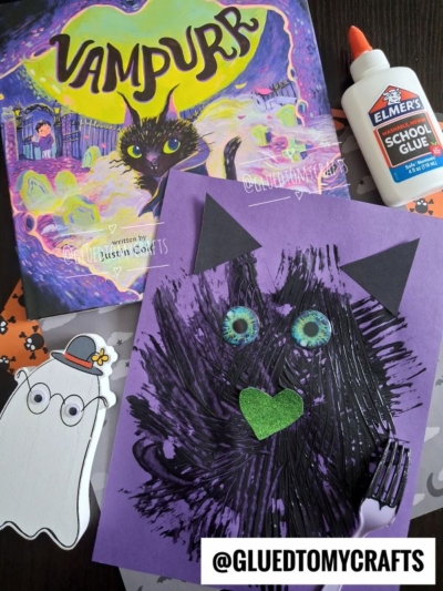 A book titled Vampurr, a fork painted cat craft featuring a purple cat with green eyes and a heart nose, glue, paper ghost, and a @GLUEDTOMYCRAFTS watermark are shown on a dark surface.