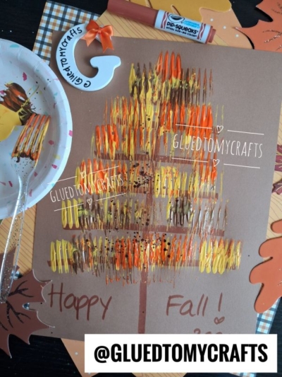 Child’s fall craft on brown paper featuring a tree trunk with colorful fork-painted leaves in yellow, orange, and brown, surrounded by a paint-covered fork, marker, paper leaves, and craft decorations.