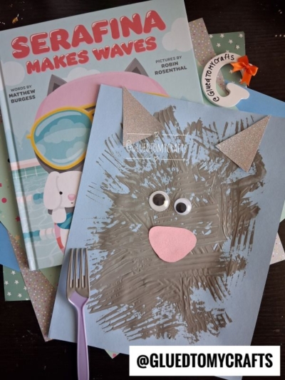 Childrens book Serafina Makes Waves next to a blue paper wolf craft made with brown paint, glittery ears, a pink nose, googly eyes, and a plastic fork used for the fur texture—perfect inspiration for your own fork painted cat or other animal creations. @GLUEDTOMYCRAFTS tag shown.