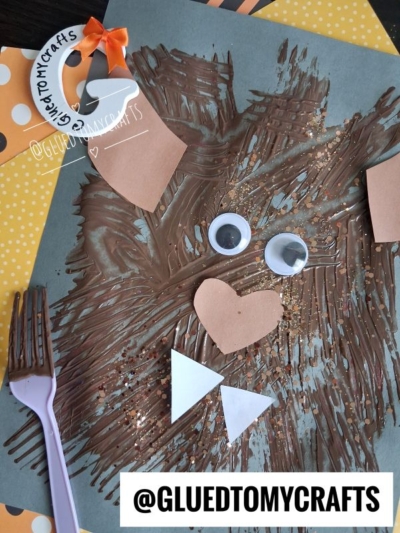 A childrens lion craft made with brown paint, googly eyes, heart and triangle paper cutouts for the face, and a purple fork. GLUEDTOMYCRAFTS is written on the image.