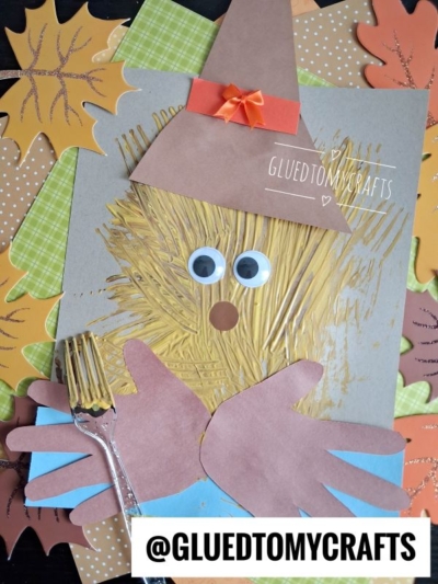 A scarecrow face craft made with a painted fork, googly eyes, paper hat, nose, hands, and surrounded by colorful paper leaves—this is one of the cutest FALL CRAFTS FOR KIDS. The handle @GLUEDTOMYCRAFTS is at the bottom.