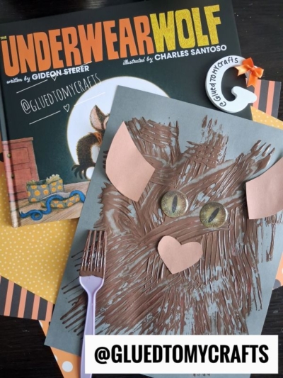 Childrens book Underwearwolf with a brown painted wolf craft, paper heart nose, circle eyes, fork, and @gluedtomycrafts sign on a table with polka dot paper and a bat-shaped ornament.