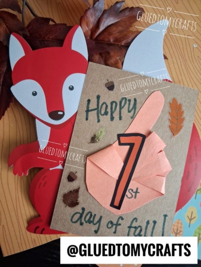 A handmade 1st day of fall card with a red fox cutout, paper leaves, and a large orange finger pointing at the number 1, laying on a fall-themed background—perfect for FALL CRAFTS FOR KIDS. @GLUEDTOMYCRAFTS is printed at the bottom.