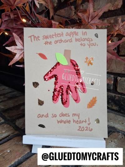 A handmade card with a red sequin apple and a brown paper stem. It reads, The sweetest apple in the orchard belongs to you, and so does my whole heart! 2026. Autumn leaves decorate the background, making it a charming example of FALL CRAFTS FOR KIDS.
