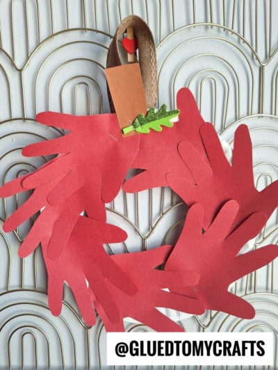 A wreath made of red paper hand shapes and a brown ribbon for hanging, decorated with a small green paper leaf and brown pot—perfect for FALL CRAFTS FOR KIDS. @GLUEDTOMYCRAFTS is written at the bottom.