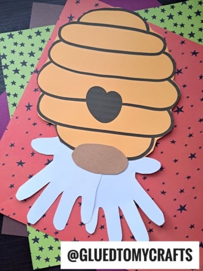 A paper craft honey bee hive with a heart-shaped entrance and cut-out hand shapes below, placed on star-patterned papers—this whimsical scene resembles a charming Paper Beehive Gnome. @GLUEDTOMYCRAFTS is written at the bottom.