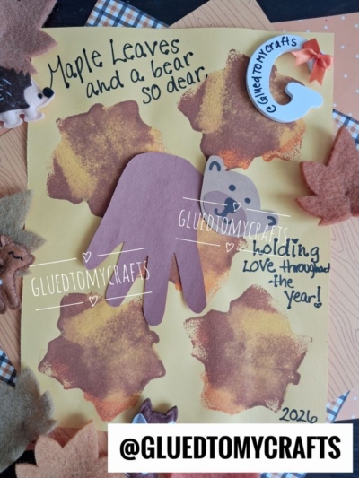 A handmade craft features a brown bear holding a maple leaf, surrounded by leaf prints. Perfect for FALL CRAFTS FOR KIDS, the text reads: Maple Leaves and a bear so dear, and holding love throughout the year! with the handle @gluedtomycrafts.