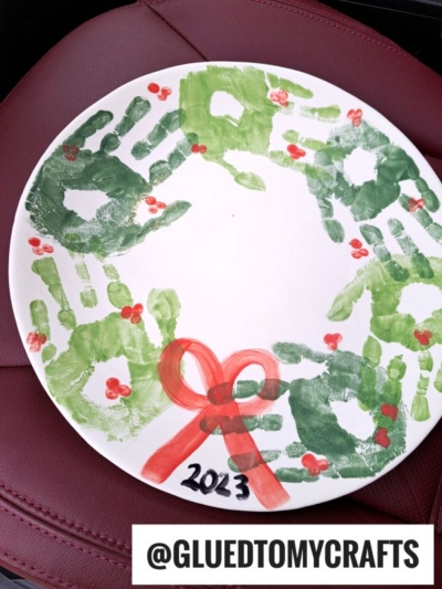 A white plate with green handprints forming a wreath, red fingerprints as berries, a painted red bow, and 2023 written in black. The plate sits on a red surface. @GLUEDTOMYCRAFTS watermark at the bottom.