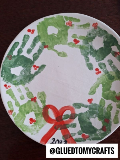 A festive paper plate wreath made with green handprints, red berry dots, and a red bow—perfect for CHRISTMAS CRAFTS FOR KIDS. “2023” appears beneath the bow, with @GLUEDTOMYCRAFTS labeled on the bottom right.