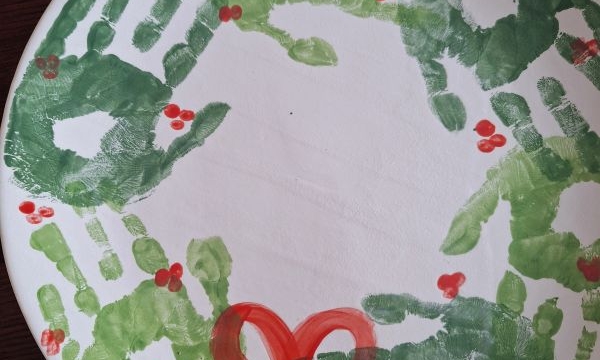 A festive paper plate wreath made with green handprints, red berry dots, and a red bow—perfect for CHRISTMAS CRAFTS FOR KIDS. “2023” appears beneath the bow, with @GLUEDTOMYCRAFTS labeled on the bottom right.