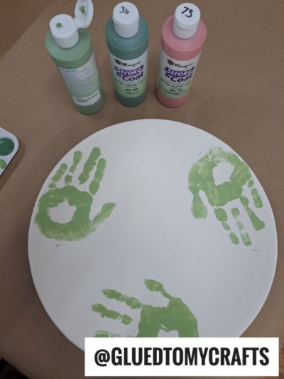 A round white surface with three green handprints, next to three bottles of green paint. A label at the bottom reads @GLUEDTOMYCRAFTS.