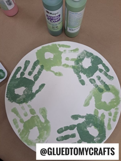 A white round board with green handprints in a circle, two bottles of green paint above it, and the text @GLUEDTOMYCRAFTS at the bottom.