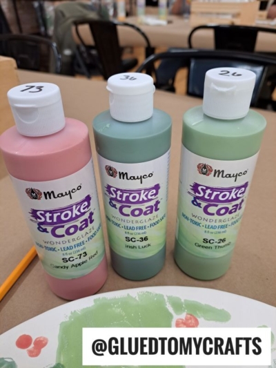 Three bottles of Mayco Stroke & Coat paint in pink (Candy Apple Red), mint green (Irish Luck), and green (Green Thumb) shades on a table with a painted plate and @GLUEDTOMYCRAFTS label in front.