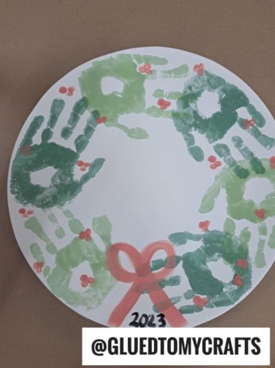 A paper plate wreath made with green handprints, red berry dots, and a red bow, labeled 2023 and @GLUEDTOMYCRAFTS at the bottom.