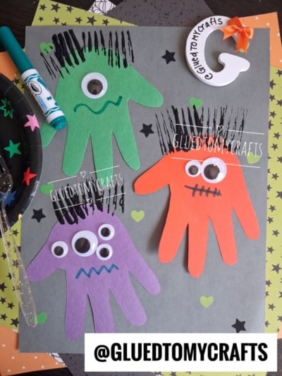 Three monster crafts made from hand-shaped paper cutouts in green, orange, and purple—each with googly eyes and marker hair—are arranged on a gray background with heart stickers and craft supplies around, making these the perfect Halloween Crafts for Kids.
