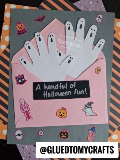 A Halloween craft shows handprint ghosts made from white paper on a pink envelope, surrounded by Halloween stickers on striped and gray paper. Perfect for anyone searching for creative Halloween Crafts for Kids, the text reads A handful of Halloween fun! and @GLUEDTOMYCRAFTS.