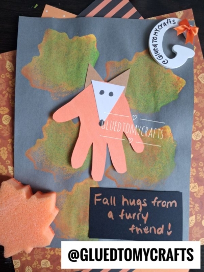 A handmade fall craft featuring a fox made from a handprint on gray paper, orange leaves in the background, a felt leaf, and the text Fall hugs from a furry friend! and @GLUEDTOMYCRAFTS—perfect inspiration for FALL CRAFTS FOR KIDS.