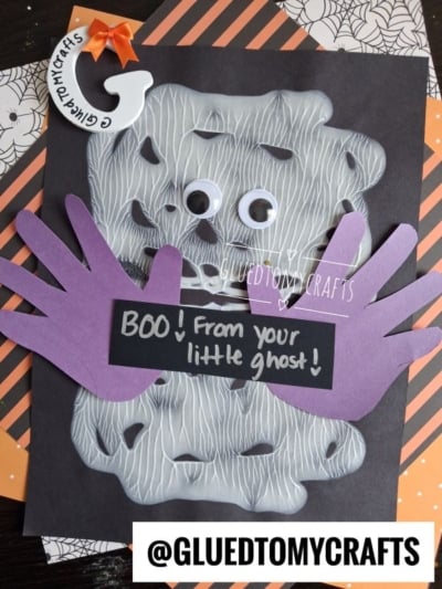 A festive Halloween craft featuring a paint splat ghost made of white paint and googly eyes, with purple handprints for wings, set against a chalkboard saying BOO! From your little ghost! on black and orange patterned paper.