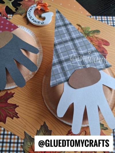 Paper plate crafts of gnomes with hand-shaped cutouts for beards, plaid paper hats, and fall leaves on an orange striped background. @GLUEDTOMYCRAFTS is written in bold at the bottom.