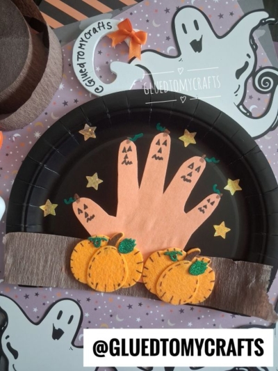 A Halloween craft featuring a handprint ghost on a black paper plate, two felt pumpkins, yellow stars, and ghost decorations on a purple background with @GLUETOMYCRAFTS text.
