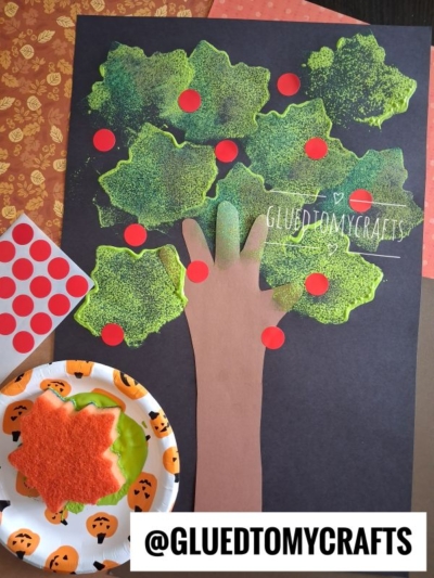 A handmade tree craft perfect for FALL CRAFTS FOR KIDS, featuring a brown paper trunk and leafy green top decorated with red dot stickers. A paper plate with pumpkin designs holds a red felt leaf and a green object. @GLUEDTOMYCRAFTS is written at the bottom.