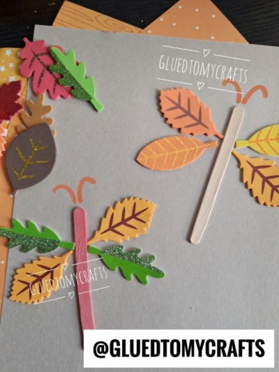 Colorful leaf and popsicle stick butterflies, featuring foam leaf sticker crafts, are arranged on a gray surface with paper leaves and autumn-themed decorations. The words GLUEDTOMYCRAFTS and @GLUEDTOMYCRAFTS appear on the image.