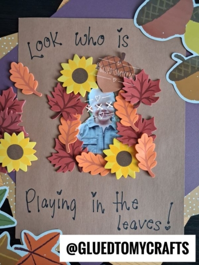 A fall-themed craft with a photo surrounded by paper leaves, sunflowers, and foam leaf sticker crafts. Text says, Look who is playing in the leaves! and @GLUEDTOMYCRAFTS is at the bottom.