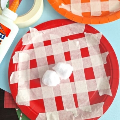 Overhead view of a kids’ craft project with red and orange paper plates decorated with masking tape in a plaid pattern, cotton balls in the center, and glue and tape placed nearby on a blue background.