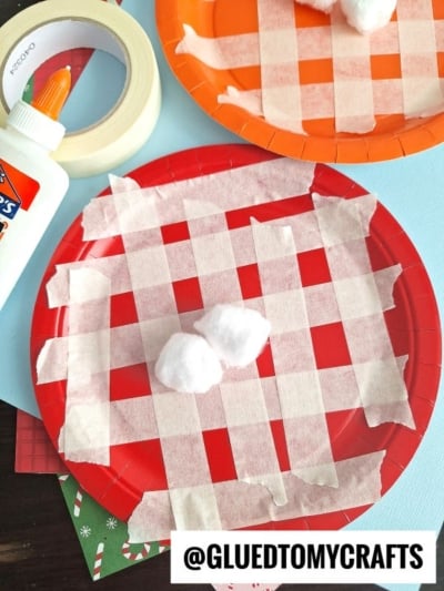 Overhead view of a kids’ craft project with red and orange paper plates decorated with masking tape in a plaid pattern, cotton balls in the center, and glue and tape placed nearby on a blue background.