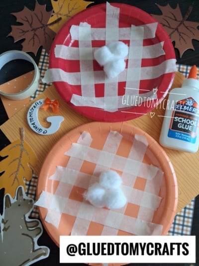Red and orange plates with masking tape grid patterns, topped with cotton balls, create a fun masking tape pie craft. Surrounded by a glue bottle, paper leaves, tape, and fall-themed crafts. @GLUEDTOMYCRAFTS is written at the bottom.