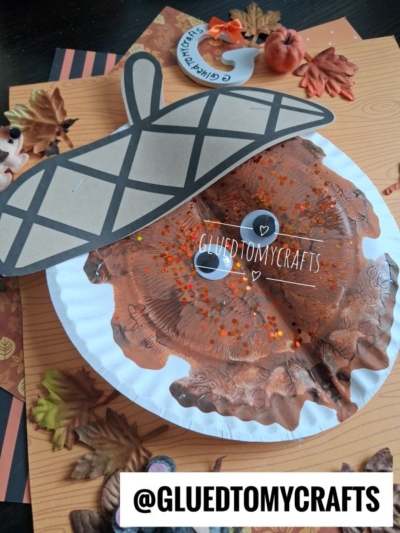 Paper plate craft shaped like an acorn, painted brown with orange glitter, large googly eyes, and a paper topper. Surrounded by fake autumn leaves and small pumpkins. @GLUEDTOMYCRAFTS text at the bottom.