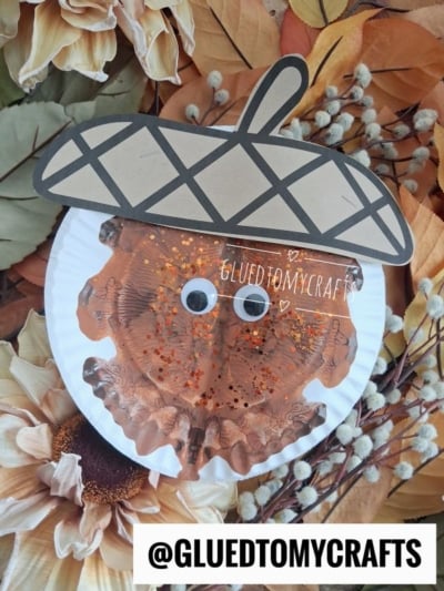A paper plate paint splat acorn craft with googly eyes, brown paint, and glitter, surrounded by artificial sunflowers and leaves. The text @GLUEDTOMYCRAFTS is at the bottom of the image.