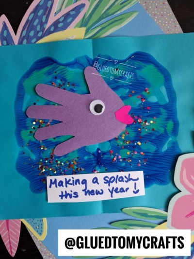 A colorful handprint fish PAINT SPLAT CRAFT with a googly eye and pink lips, surrounded by painted blue water and glitter. Text below reads, Making a splash this new year! with @GLUEDTOMYCRAFTS at the bottom.